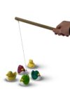 Eine Hand hält einen Holzstab an dem ein Faden mit Magnet ist. Des Weiteren sind 5 bunte Kunststoffenten zu sehen.