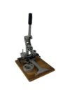 Eine manuelle Buttonmaschine aus Metall mit langem Hebelgriff, montiert auf einer rechteckigen Holzplatte. Die Maschine verfügt über mehrere runde Aufnahmen und mechanische Komponente.