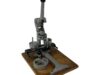 Eine manuelle Buttonmaschine aus Metall mit langem Hebelgriff, montiert auf einer rechteckigen Holzplatte. Die Maschine verfügt über mehrere runde Aufnahmen und mechanische Komponente.