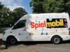 Ein großes weißes Fahrzeug mit bunter Aufschrift "Spielmobil" und bunten Stickern die Roller, Springseile, Diabolos, Hulahoops, Bälle, Bastelmaterial und Handabdrücke zeigen
